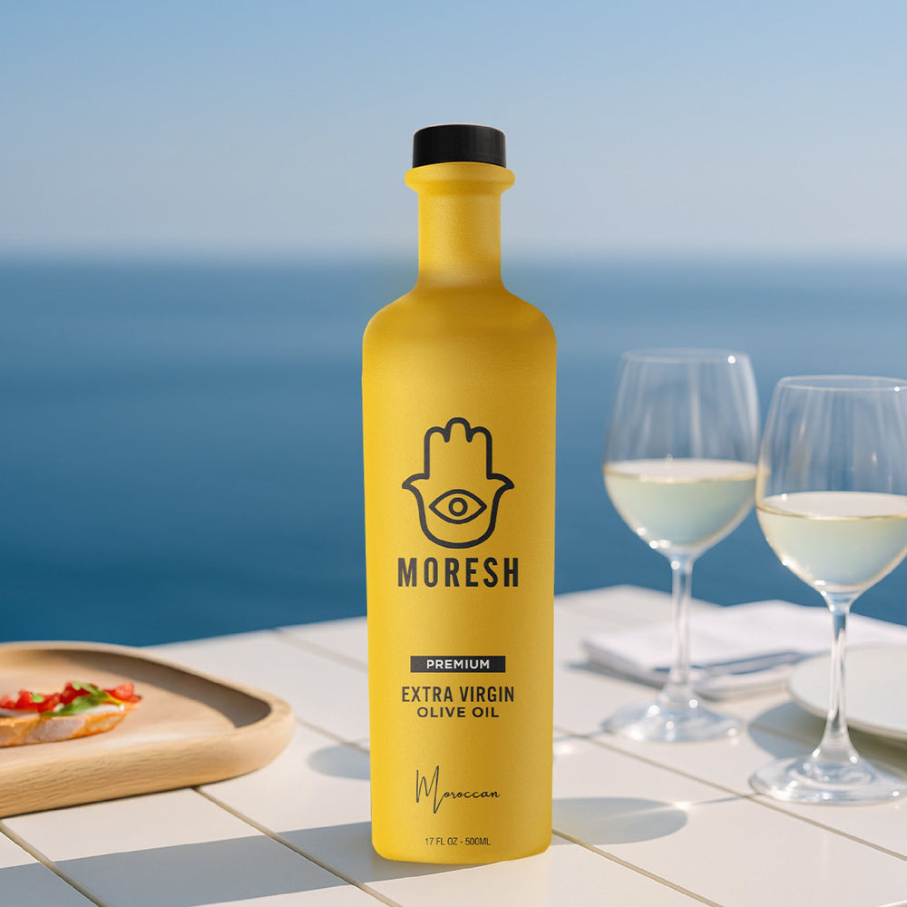 
                  
                    Yellow bottle of Moresh olive oil with two glasses of white wine and a plate of food on a table by the ocean.
                  
                